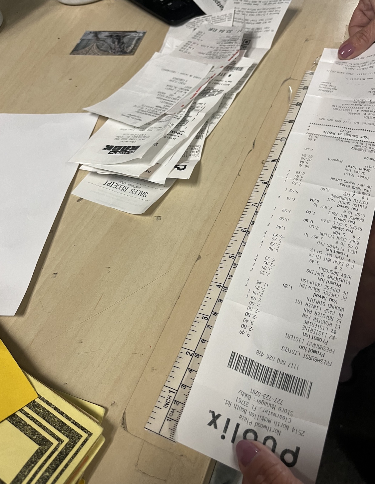 A long grocery receipt held against a 12-inch ruler, stretching well past its full length, on a wooden desk with other receipts